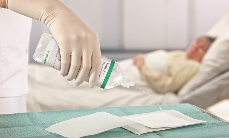 V popredí ruka v lekárskej rukavici nalieva irigačný roztok Prontosan na podložku ležiacu na stole vedľa tuby  Askina Calgitrol. V pozadí leží pacientka na nemocničnom lôžku.