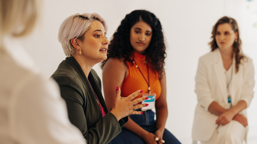 Štyri ženy sedia vedľa seba na mentoringovom programe pre ženy. Cieľom je zviditeľniť ženy v organizácii a pomôcť im v ich osobnom raste. Druhá žena pozoruje prvú, sivovlasú ženu v zelenom saku, pričom hovorí a dáva znamenie rukou.  