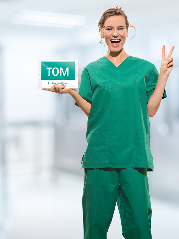 Zdravotníčka v lekárskom oblečení drží tablet so skratkou „TOM“. Prstami tiež ukazuje znak „V“